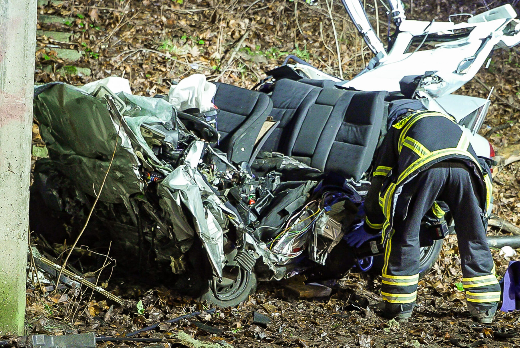 Ein Feuerwehrmann arbeitet in der Nacht an einem völlig zerstörten Mercedes, der frontal gegen ein Brückenbauwerk geprallt ist. Das Fahrzeug ist stark deformiert, Wrackteile liegen auf der Fahrbahn.