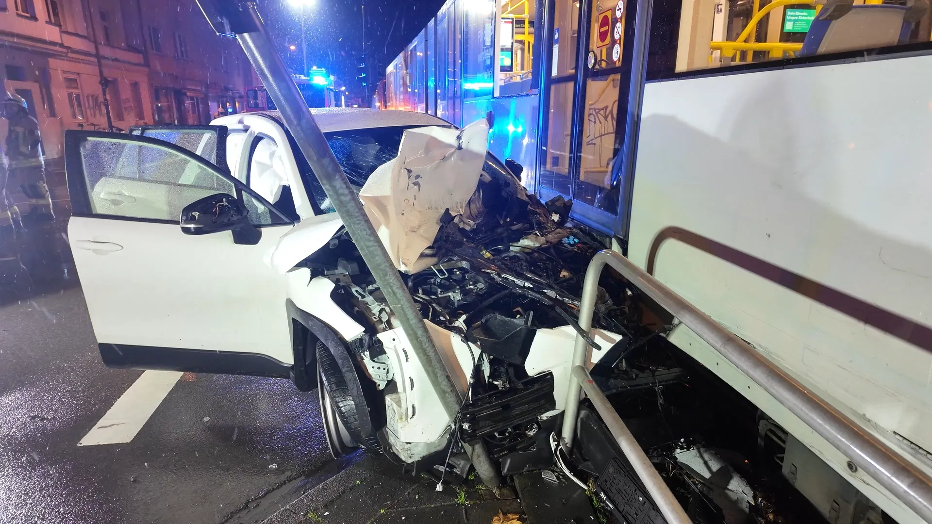 Schwer beschädigter weißer Mietwagen steht nach einer Kollision dicht an einer Straßenbahn der KVB-Linie 13 in Köln-Sülz. Die Front des Autos ist stark eingedrückt, Airbags sind ausgelöst, Feuerwehrkräfte arbeiten im Hintergrund an der Unfallstelle.