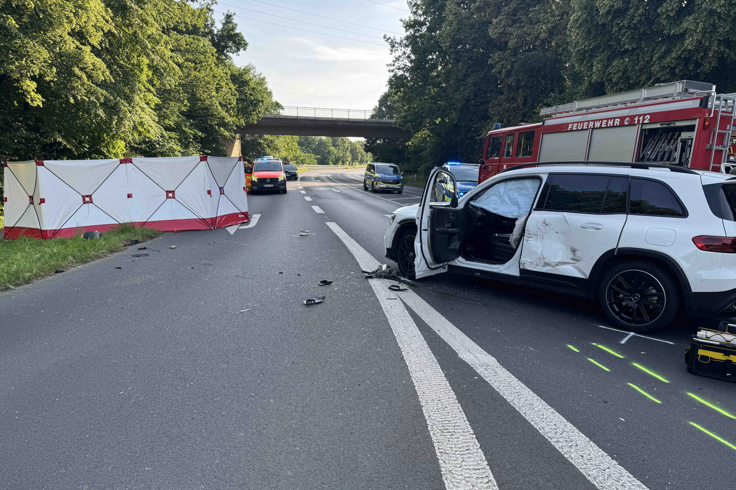 Unfallstelle auf der B477 in Bergheim mit beschädigtem weißen SUV, Sichtschutz und mehreren Rettungsfahrzeugen vor einer Straßenüberführung.